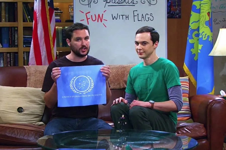 Sitcom scene of two men holding up a flag in front of a whiteboard reading 'FUN WITH FLAGS.'