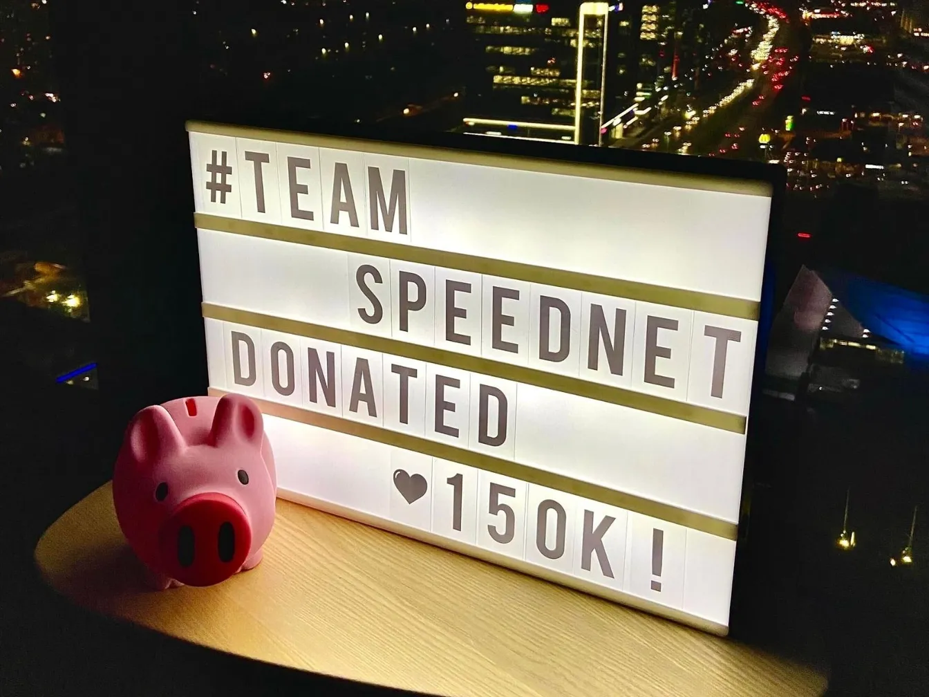 Świecący napis "#TEAM SPEEDNET DONATED ♥150K!" obok skarbonki w kształcie świnki, na tle panoramy miasta nocą.