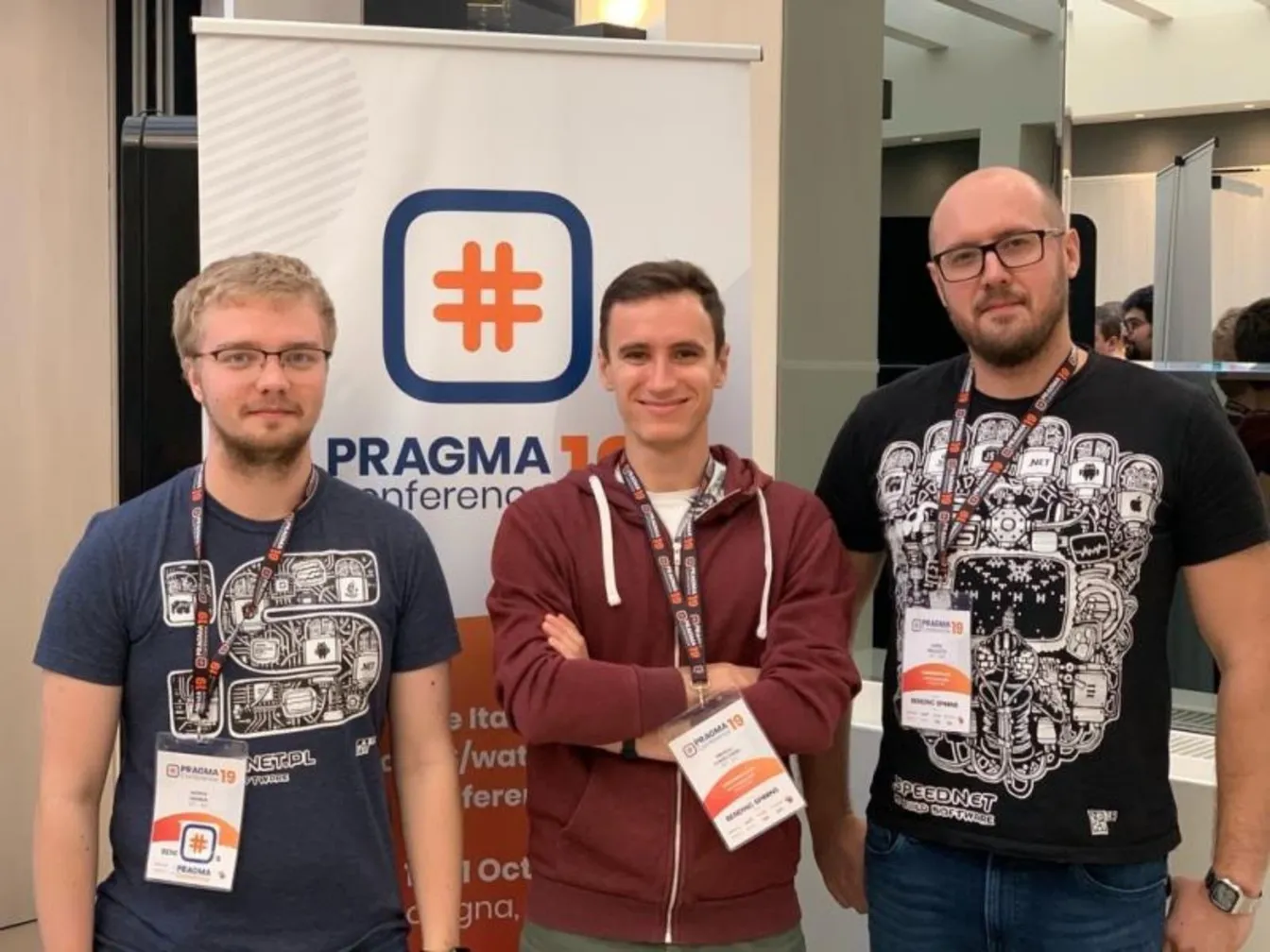 Trzech uczestników w koszulkach Speednet przed rollupem konferencji Pragma 19.