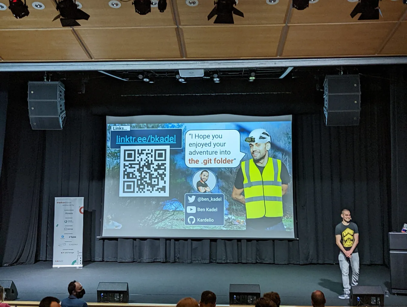 Conference speaker standing on stage beside his closing slide with contact links and a quote credited to Ben Kadel.