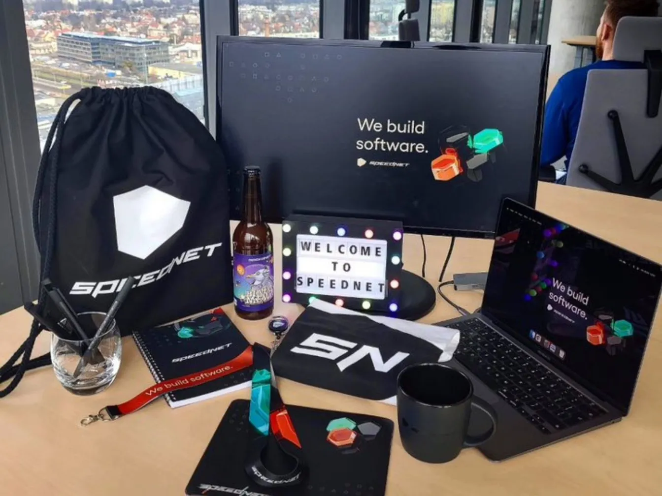 Biurko z gadżetami Speednet: torba, notes, kubek i podkładka, obok laptop z napisem 'We build software.' i świecący napis 'Welcome to Speednet'.