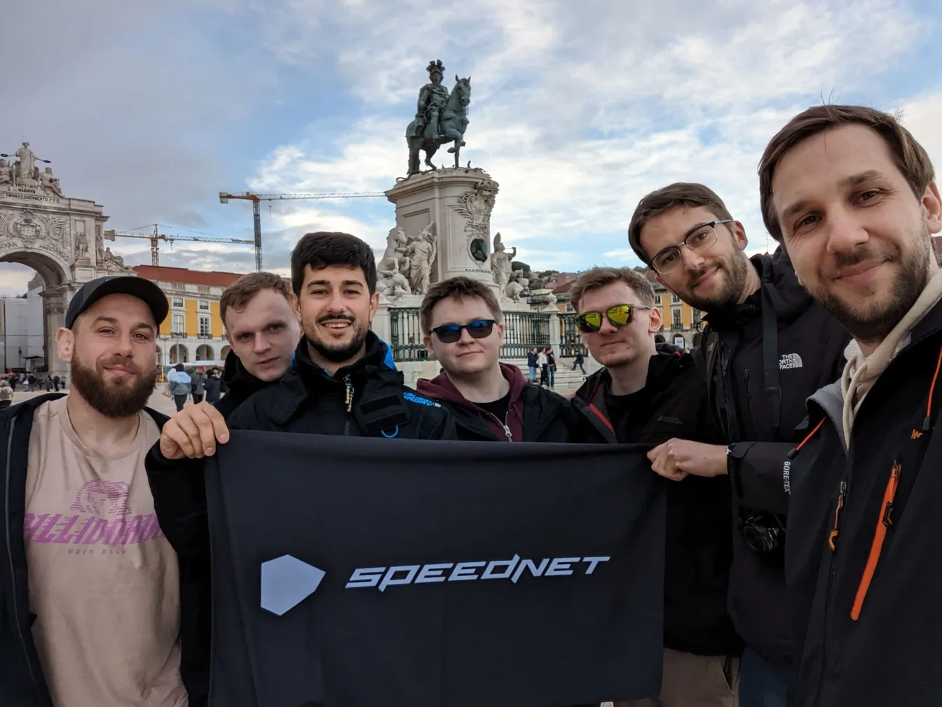 Team holding a Speednet banner in front of the equestrian statue on Lisbon's Praça do Comércio square.