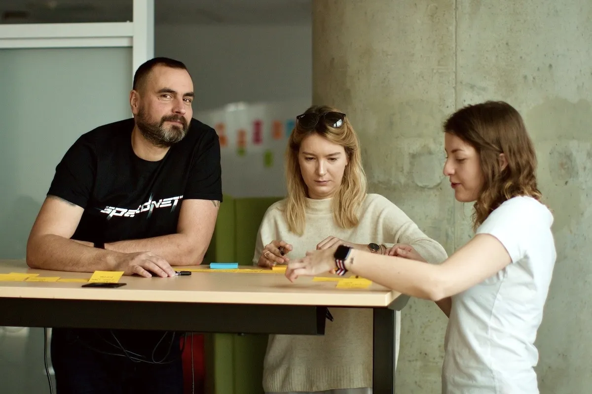 Troje pracowników rozmawia przy stole z żółtymi karteczkami, mężczyzna ma koszulkę z logo Speednet.