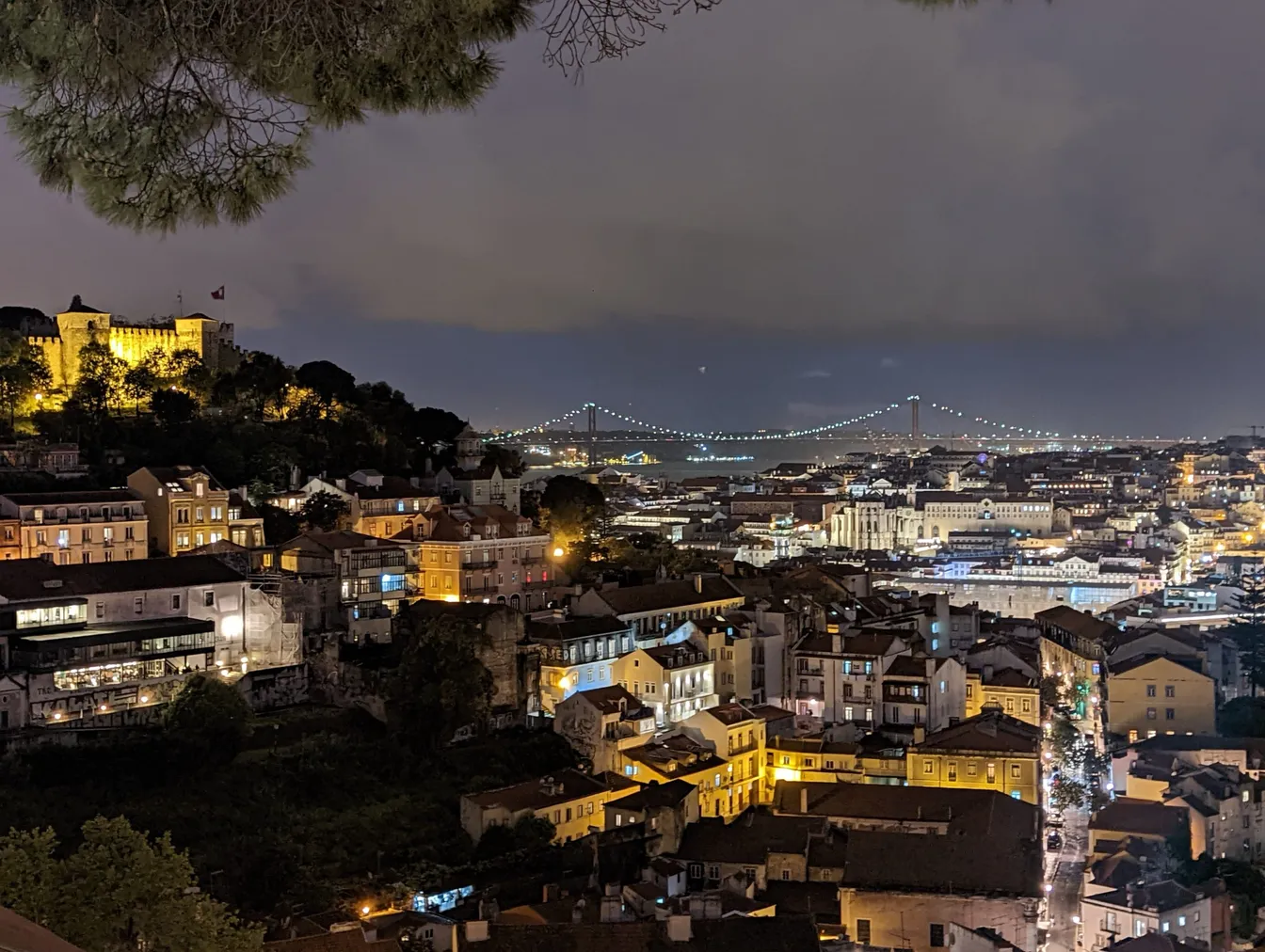 Night view over Lisbon's rooftops, with São Jorge Castle lit on the hill and the 25 de Abril bridge in the distance.