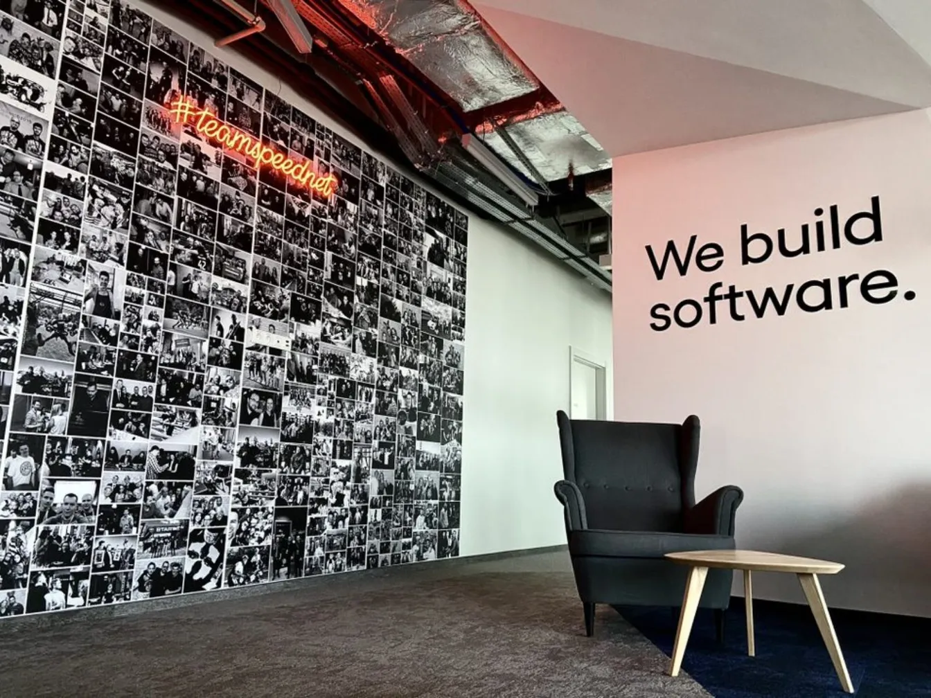 Wnętrze biura Speednet: ściana ze zdjęciami zespołu i neonowy napis '#teamspeednet', obok hasło 'We build software.' przy fotelu.