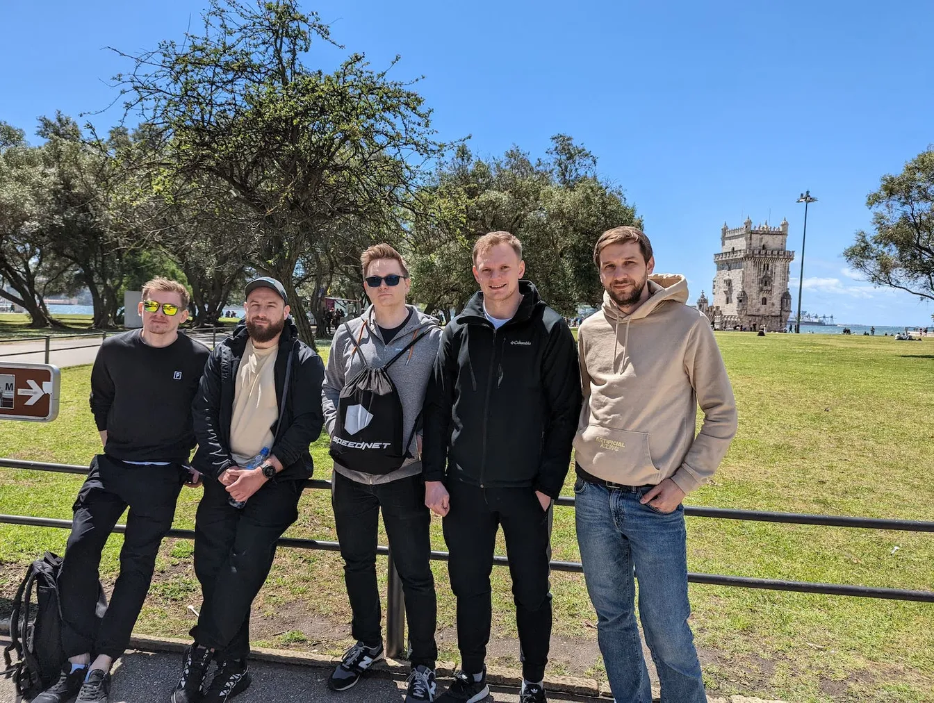 Group posing in a riverside park with Belém Tower in the background, one carrying a Speednet bag.