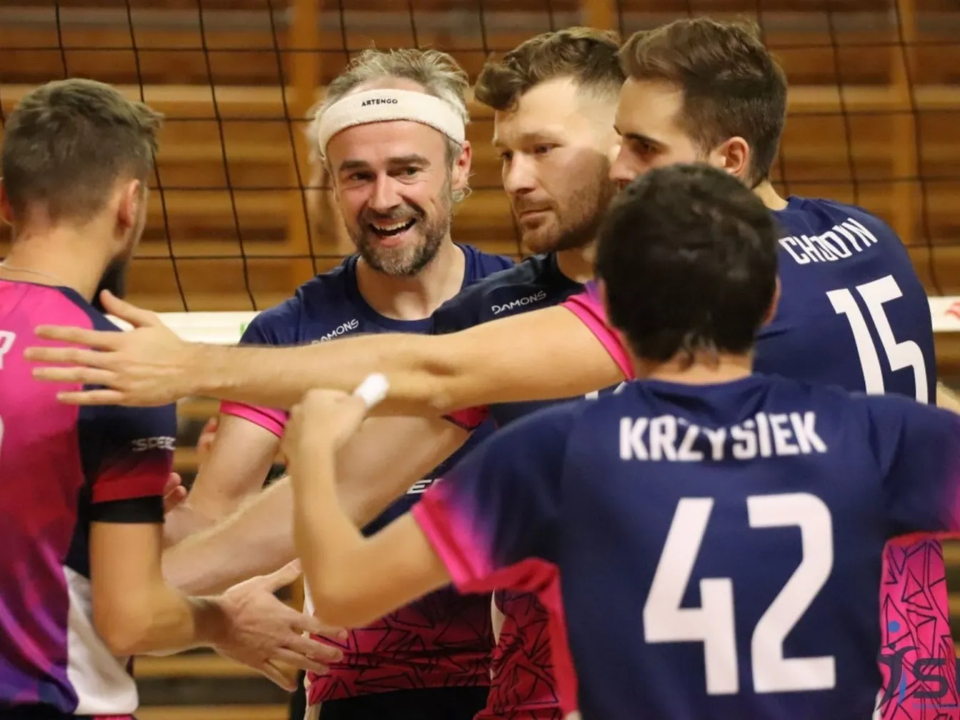 Zawodnicy siatkówki cieszą się przy siatce po zagrywce, w kolorowych strojach meczowych.