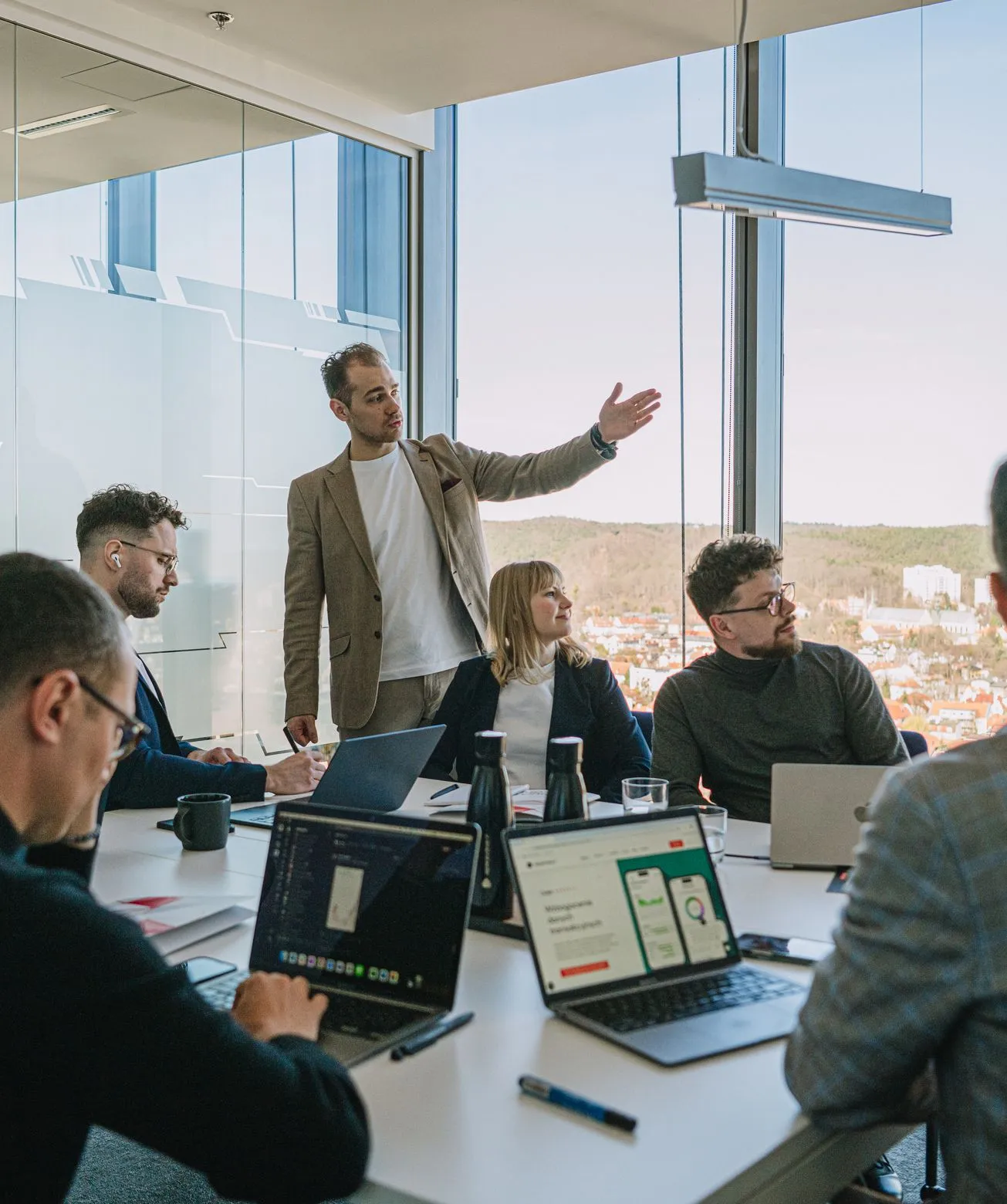 A Speednet team member presenting to colleagues at a meeting table with laptops, in a glass-walled room overlooking the city.