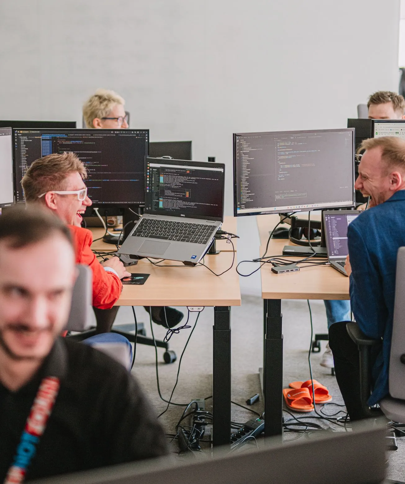 Speednet team members laughing at their desks, with monitors showing code in an open-plan office.